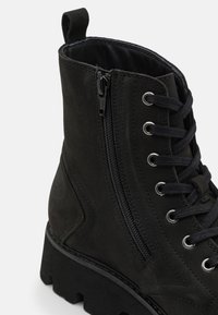 Bottine noire en daim avec une surface texturée, dotée d'une fermeture éclair sur le côté et d'un laçage à l'avant, ainsi qu'une semelle épaisse.