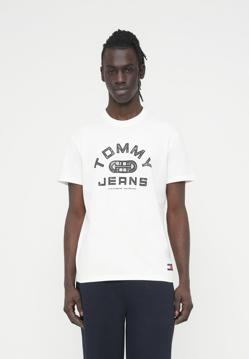 Λευκό βαμβακερό t-shirt με έντονο μαύρο λογότυπο "TOMMY JEANS". Στρογγυλή λαιμόκοψη και κοντά μανίκια, με μικρό λογότυπο σημαίας στο κάτω μέρος.