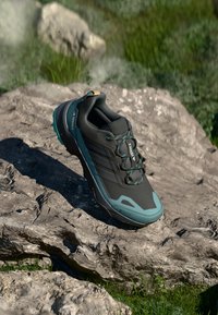 Shoe desportivo preto com detalhes em turquesa, parte superior em malha texturizada, sistema de ataque seguro, biqueira reforçada e sola robusta sobre uma superfície rochosa.