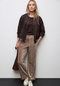 Hnědá bomber jacket s žebrovaným límcem, tmavě hnědý top, béžové široké kordové kalhoty a bílé tenisky. Stojí proti neutrální stěně.