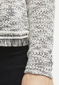 Gros plan sur la manche d'un pull en tricot gris et blanc avec un détail de franges au bas, porté sur un pantalon noir sur le bras d'une personne.