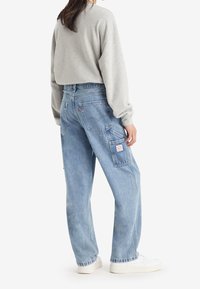 Jeans de denim azul claro con un corte relajado, que presentan múltiples bolsillos, una etiqueta de parche, y se combinan con una camiseta de manga larga gris y zapatillas blancas.