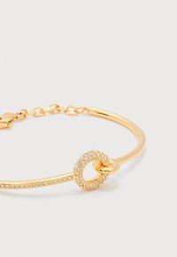 Χρυσό βραχιόλι με κυκλικό γούρι διακοσμημένο με μικρά διαφανή κρυστάλλινα στοιχεία και γυαλιστερό χρυσό κρίκο που περνάει μέσα από το γούρι, σε λευκό φόντο.