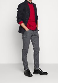 Man i svart kavaj över röd tröja, grå jeans och svarta klädsko med händerna i fickorna mot en enfärgad bakgrund.