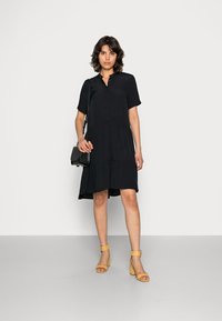 Vestido preto em camadas com mangas curtas e colarinho abotoado. Combinado com sandálias amarelas de salto bloco e uma pequena bolsa crossbody preta.