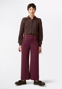 Schwarzbluse mit floralem Muster, langen Ärmeln und geknöpften Manschetten, kombiniert mit bordeauxfarbenen High-Waist-Hosen mit weitem Bein und goldenen Knöpfen.