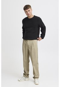 Pull noir en maille avec encolure ronde, associé à un pantalon beige ample. Baskets blanches avec des accents bleu marine. Fond neutre.