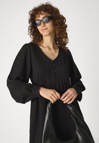 Vestido preto de mangas longas com frente abotoada, textura plissada e silhueta evasê; combinado com óculos de sol oversized pretos e uma bolsa preta.