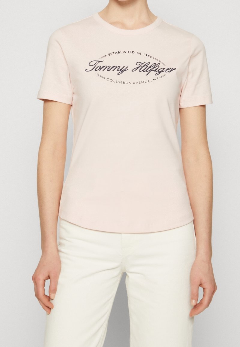 Světle růžové tričko s krátkým rukávem vyrobené z měkké bavlny, s tmavým tištěným logem "Tommy Hilfiger" na hrudi.