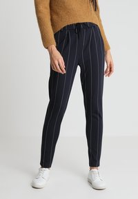 Navy trousers with vertical white stripes, drawstring waist, tapered fit, and lightweight material; paired with white sneakers.