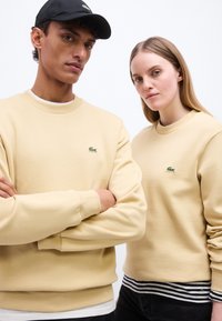 Δύο μοντέλα φορούν ίδιες μπεζ φούτερ Lacoste και στέκονται δίπλα-δίπλα σε φόντο απλό λευκό.