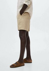 Shorts in cotone beige con taglio dritto, tasche laterali, abbinati a sandali in camoscio marrone con doppie cinghie e fibbie.