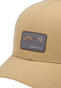 Beige cap with a flat brim and textured fabric. Features a rectangular patch with multicolored mountain and wave designs. Gray stitching details.