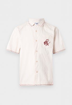 Krémová košeľa s krátkym rukávom s červeným krížikovým lemom na golieri, rukávoch a spodnom leme a červenou výšivkou ruže na ľavej náprsnej vrecku.