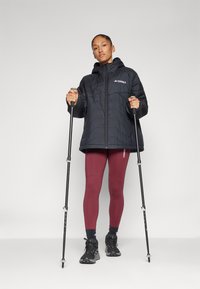 Doudoune noire avec capuche et logo, leggings bordeaux, chaussures noires et bâtons de trekking. Texture lisse, coupe ample et design en couches.