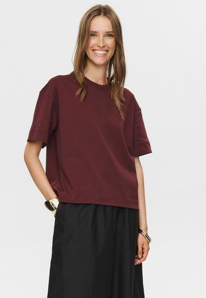 Burgunderfarbendes T-Shirt mit kurzen Ärmeln in lockerer Passform, aus weicher Baumwolle, mit Rundhalsausschnitt und sauberem Saum. Kombiniert mit schwarzen Hosen.