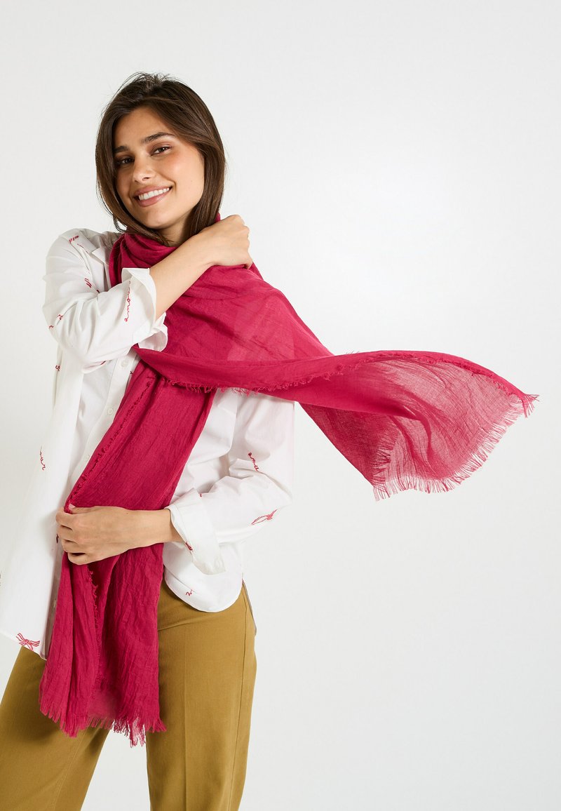 Écharpe légère fuchsia en tissu doux, avec des bords effilochés, drapée de manière décontractée sur les épaules, associée à une chemise blanche et un pantalon beige.