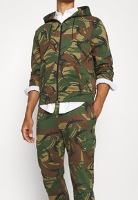 Grön och brun kamouflage zip-up hoodie med dragsko huva, med en mjuk textur och sidofickor, kombinerad med matchande byxor.