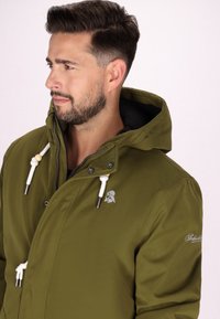 Giacca verde oliva con cappuccio, dotata di cordini bianchi, componenti in metallo argentato e un logo sul petto. Texture liscia e vestibilità sartoriale.