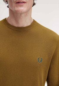 Maglione a maglia giallo senape con scollo rotondo e motivo testurizzato, con un piccolo logo ricamato blu sul petto.