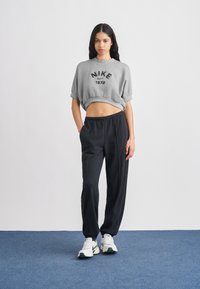 Γκρι κοντομάνικο φούτερ με μαύρο τύπωμα "NIKE 1972", μαύρο φαρδύ παντελόνι και λευκά αθλητικά παπούτσια με πράσινες λεπτομέρειες· φόντο επίπεδο.