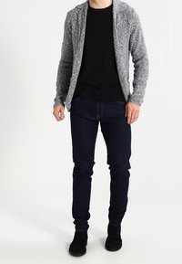 Cardigan grigio a maglia con apertura frontale, t-shirt nera, jeans scuri e stivaletti neri alla caviglia. Texture liscia e design aderente.