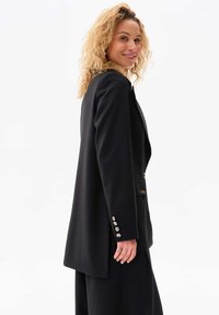 Zwarte blazer met een getailleerde pasvorm, sluiting met één knoop en zilveren knoopaccenten op de mouwen. Gladde textuur en scherpe ontwerp.