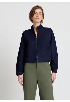 Femme portant une veste zippée bleu marine à col montant et un pantalon taille haute vert olive, debout devant un fond clair uni.