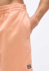 Κοντινό πλάνο προσώπου που φοράει ροδακινί σορτς με ελαστική μέση, με το χέρι στην πλαϊνή τσέπη και μικρό μαύρο και λευκό λογότυπο κοντά στο τελείωμα.