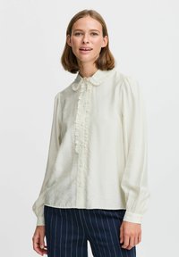 Helle Creme-Bluse mit Rüschkragen und Knopfleiste. Lange, puffige Ärmel mit Knopfmanschetten. Weicher, glatter Stoff.