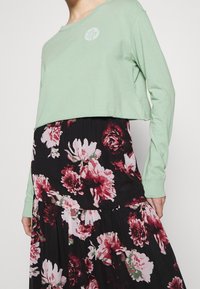 Zelený krátký top s dlouhými rukávy a bílým logem, v kombinaci s černou květinovou sukní s růžovými a bílými květy a vrstveným designem.