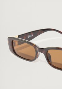 Brunefargede solbriller med ramme i skilpadde-mønster. Firkantet design med en subtil gullakkent ved stangen. Logotype synlig.