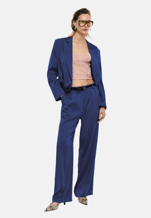 Donna che indossa un completo blu oversize con crop top in pizzo rosa, grandi orecchini, occhiali con lenti gialle e tacchi a punta con motivo serpente, in piedi con sicurezza.