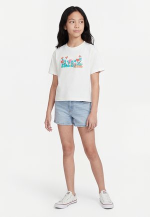 Mädchen mit langen schwarzen Haaren, das ein weißes T-Shirt mit Blumenmuster, helle Jeansshorts und weiße Turnschuhe trägt und vor weißem Hintergrund steht.