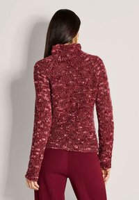 Vrouw met lang zwart haar, gekleed in een bordeauxrode en roze grove gebreide coltrui en bijpassende bordeauxrode broek, met de rug naar ons toe.
