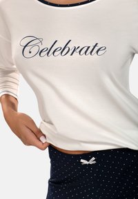 Krém színű felső "Celebrate" felirattal, tengerész kék pöttyös rövidnadrággal és ezüst szalag részlettel. Puha anyag, laza fazon, kerek nyakkivágás és háromnegyedes ujj. 