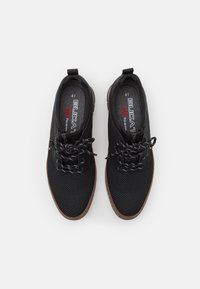 Black sneakers with a textured upper, black laces, and brown rubber soles. Features branding on the insoles and a pull tab at the heel.
