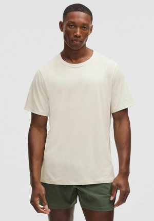 Hellbeige T-Shirt mit Rundhalsausschnitt und kurzen Ärmeln. Glatte Textur, lockere Passform. Kombiniert mit dunkelgrünen Shorts, ohne Muster oder sichtbare Hardware.
