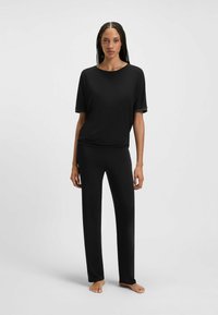 Vrouw die blootsvoets staat in een eenvoudige zwarte t-shirt met korte mouwen en bijpassende zwarte rechte broek, op een effen lichtgrijze achtergrond.