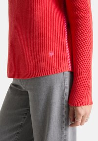 Roter gerippter Pullover mit langen Ärmeln, ausgestattet mit einem seitlichen Schlitz und kleinem rosa Herz-Logo am Saum, kombiniert mit grauen Jeans.