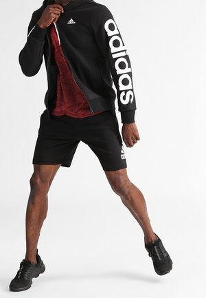 Man wearing black Adidas hoodie and shorts with white logo, mid-action with legs apart and one hand gripping the hoodie zipper.