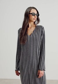 Robe longue à manches longues noir et argent à rayures avec un décolleté en V et un tissu texturé. Le mannequin porte des lunettes de soleil noires, se tenant devant un fond uni.