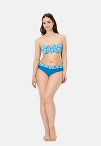 Joven de pie descalza, vistiendo un bikini azul sin tirantes con patrones blancos de hojas en la parte superior y la cintura, sonriendo a la cámara.