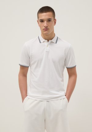Joven de pie con las manos en los bolsillos, lleva una camiseta polo blanca con cuello y bordes de las mangas a rayas negras sobre un fondo liso.