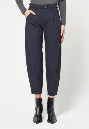 Woman wearing navy high-waisted tapered pants with pleats, a gray long-sleeve top, and black ankle boots.