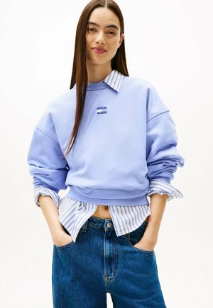 Felpa oversize azzurro chiaro con orlo e polsini a coste, indossata sopra una camicia a righe bianca e blu, abbinata a jeans di denim blu.