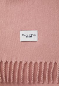 Rosa vevd teppe med vridde frynser. Har et merke med teksten "Marc O'Polo DENIM" i marineblått på hvit bakgrunn. Myk tekstur.