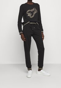 Svart sweatshirt med guldfärgade nitar i form av löv, kombinerad med svarta joggers med guldfärgade randiga muddar och dragsko i midjan, kompletterat med vita sneakers.