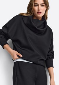 Czarna bluza z dekoltem typu cowl, z opadającymi ramionami i długimi rękawami, wykonana z miękkiego, teksturowanego materiału. W połączeniu z czarnymi spodniami.