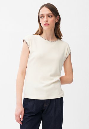 Młoda kobieta o brązowych włosach ubrana w kremowy, bezrękawnik z ciemnymi wykończeniami szwów oraz ciemne, luźne spodnie, pozująca na białym tle.